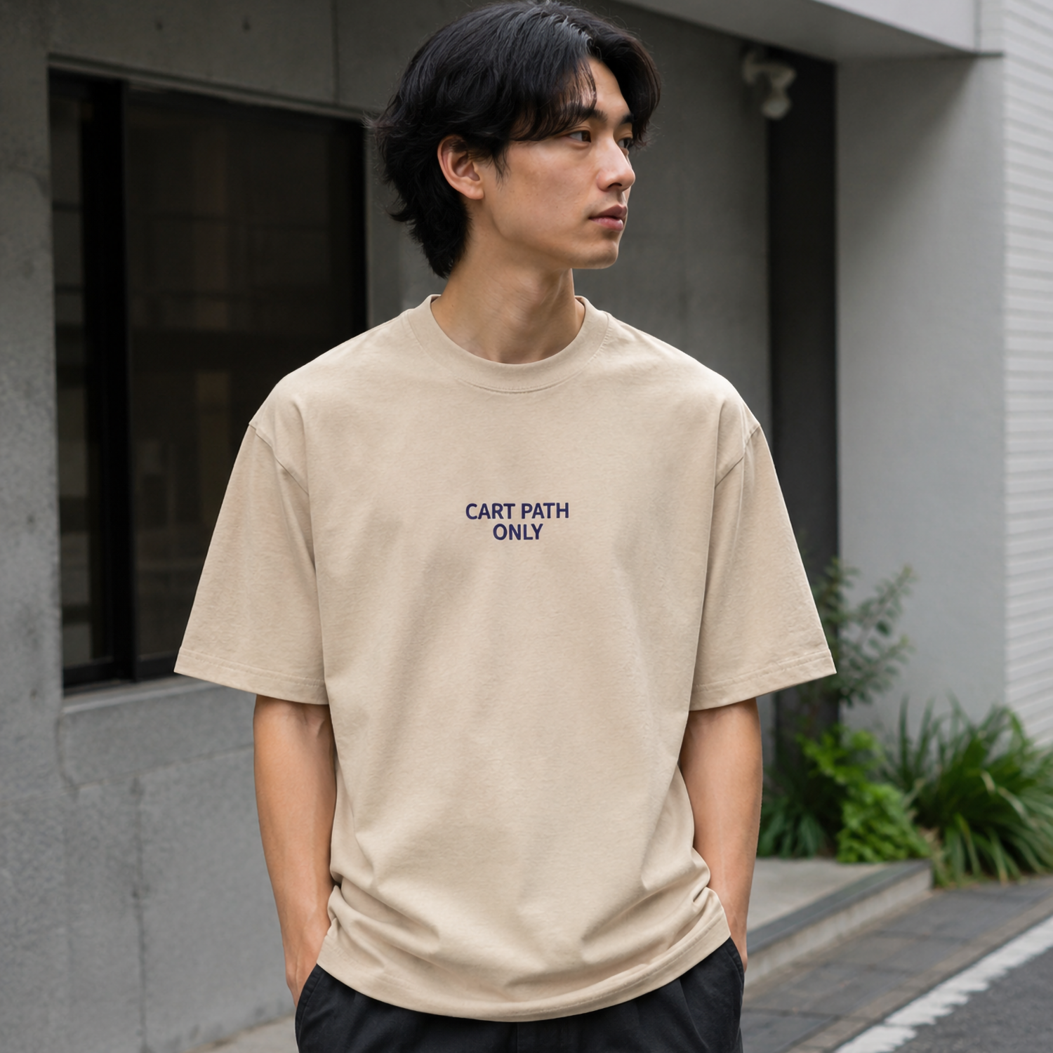 A man wearing a beige crewneck t-shirt with the text 'CART PATH ONLY' printed in navy blue on the chest, paired with black pants, standing outdoors against a gray concrete building.