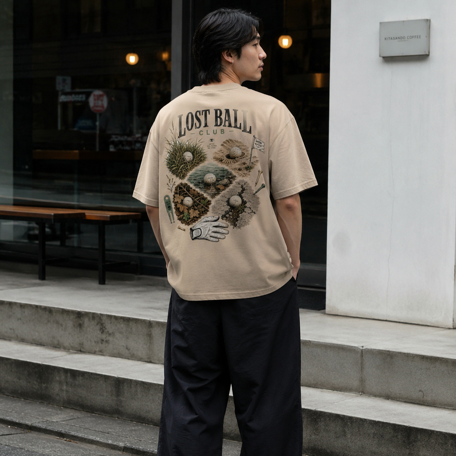 A man standing outdoors on concrete steps, viewed from behind, wearing a beige graphic t-shirt with 'LOST BALLS' and a surreal illustration on the back, paired with loose black pants and white sneakers.