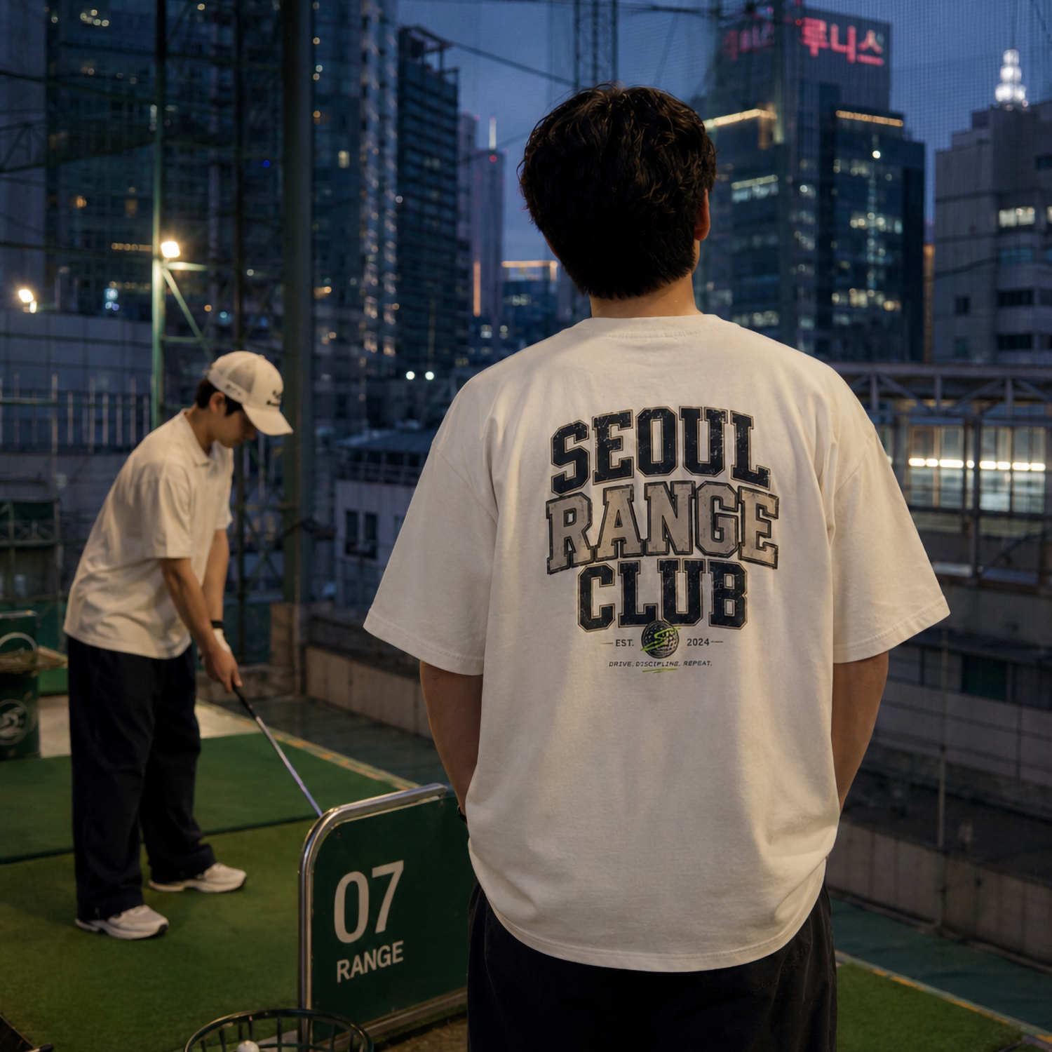 A man stands on a rooftop golf simulator at dusk, viewed from behind wearing a white t-shirt with 'SEOUL RANGE CLUB' printed in dark letters on the back. Another person is visible hitting golf balls in the background.