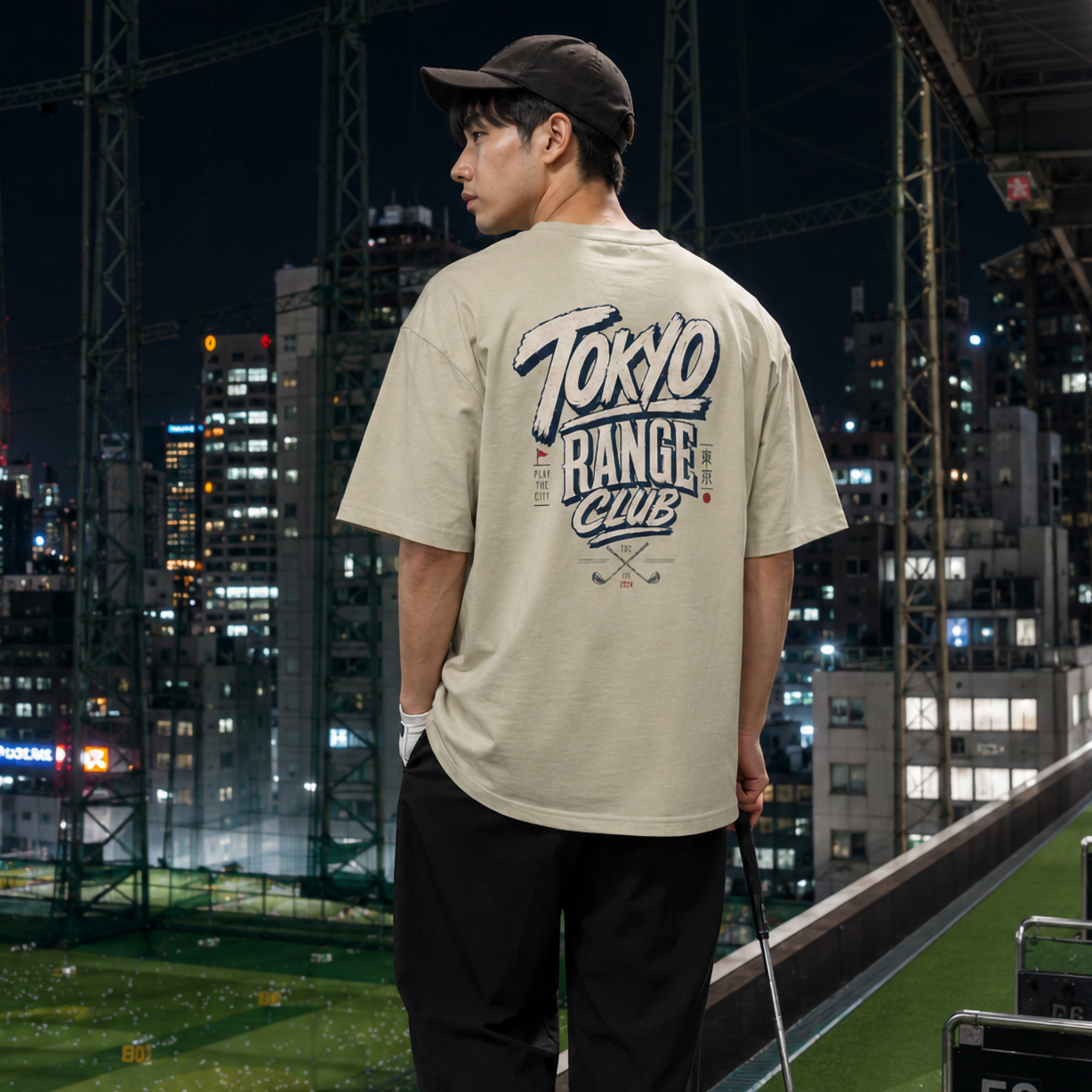 A man stands on a golf driving range at night, viewed from behind. He is wearing a beige short-sleeve t-shirt with a large 'Tokyo Ranger Club' graphic on the back, black pants, white sneakers, and a dark baseball cap.